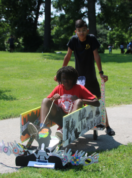 Teenager pushes another teenager in a decorated wooden go-cart.